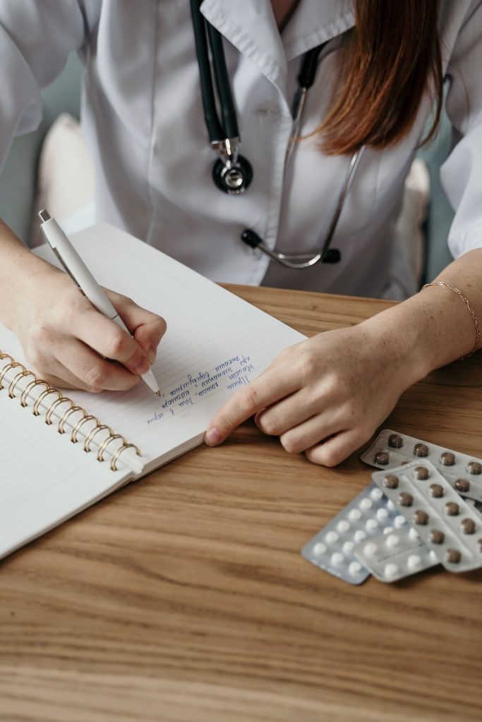 pexels-photo-5407208-5407208 Close-up of a doctor writing a prescription with medicine on a wooden desk.